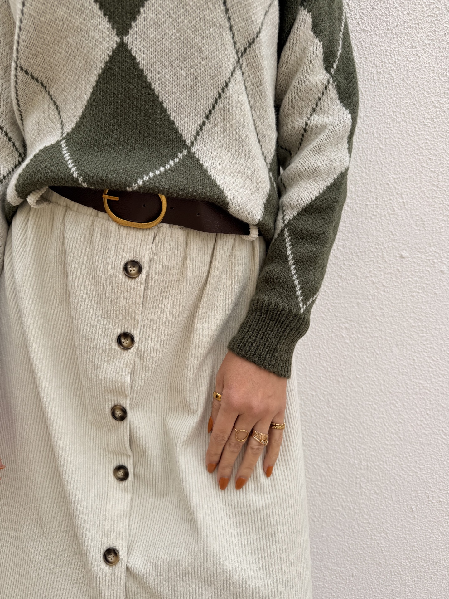 Suéter de malha verde e bege com padrão de losangos, cinto castanho com fivela dourada, saia creme com botões e mão com anéis dourados