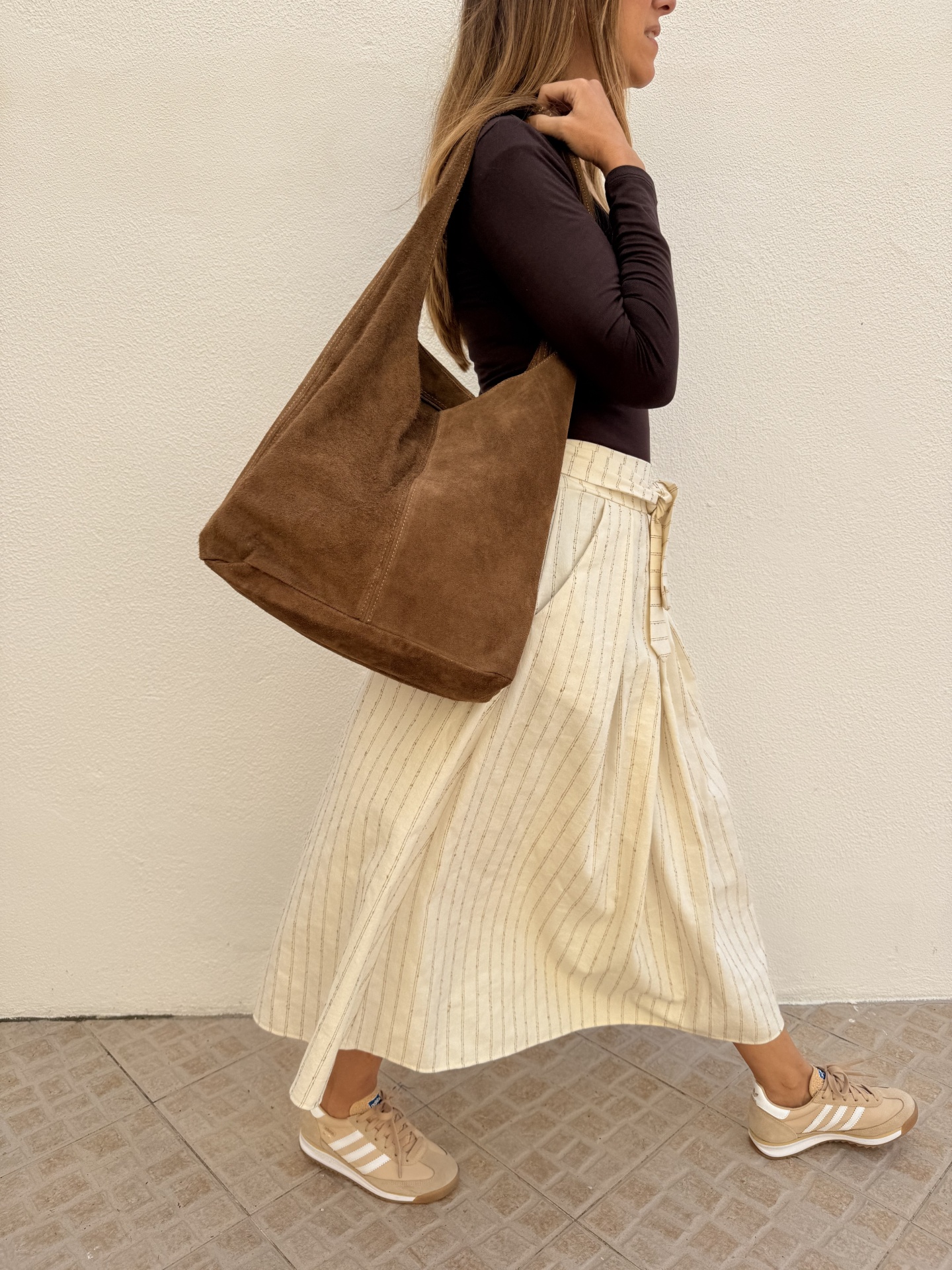 Mulher com saco castanho em tecido camurça, camisola castanha, saia creme às riscas e ténis bege com riscas brancas