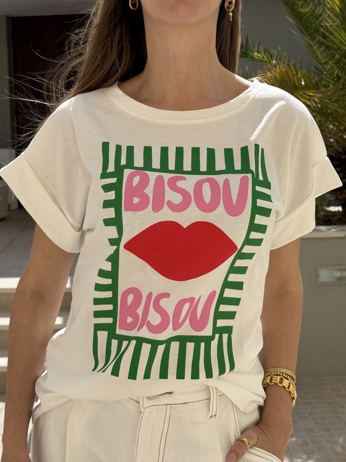 T-shirt branca com estampado colorido e texto BISOU BISOU usada por modelo com acessórios dourados.