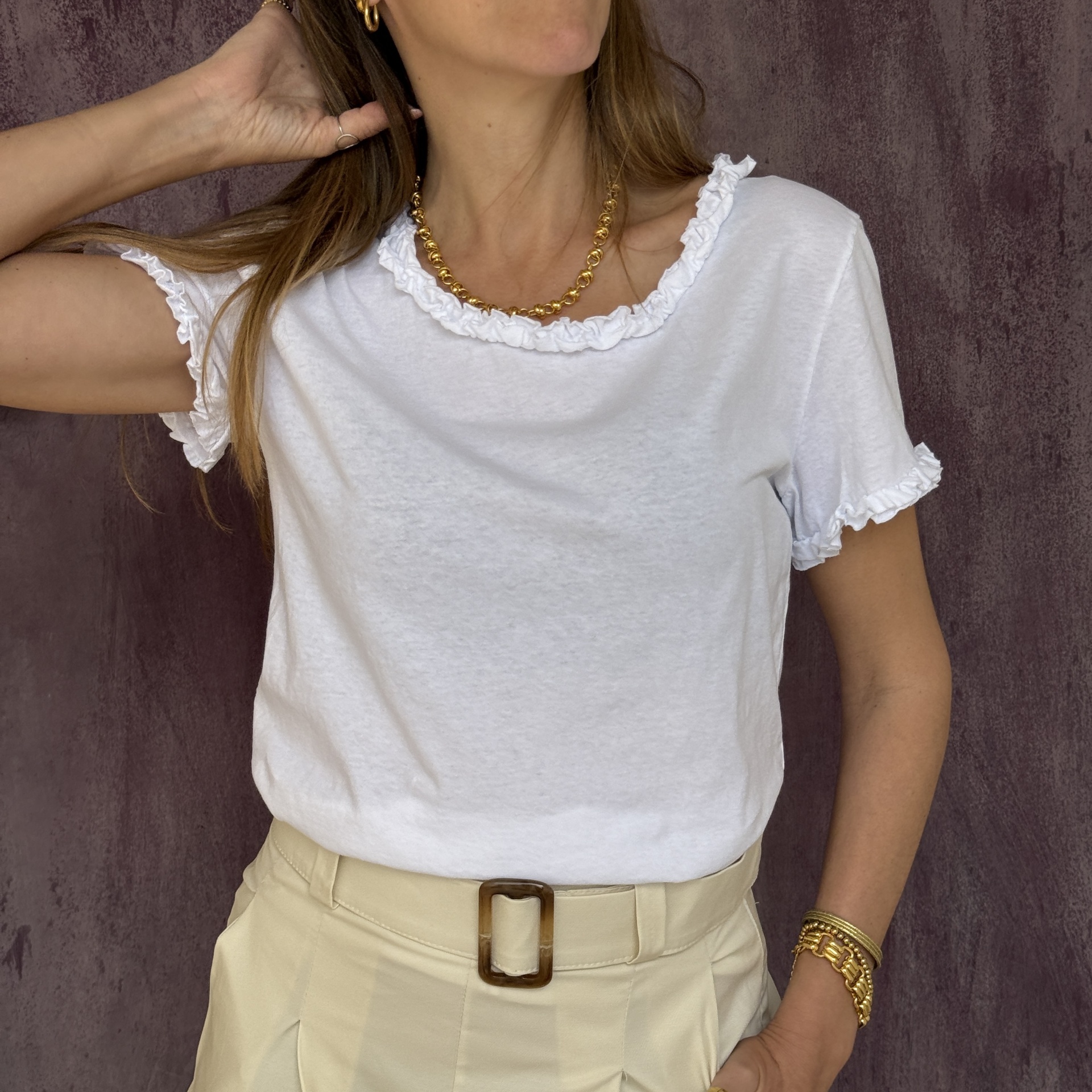 T-shirt branca com folhos nas mangas e decote, calças bege com cinto, acessórios dourados, fundo roxo