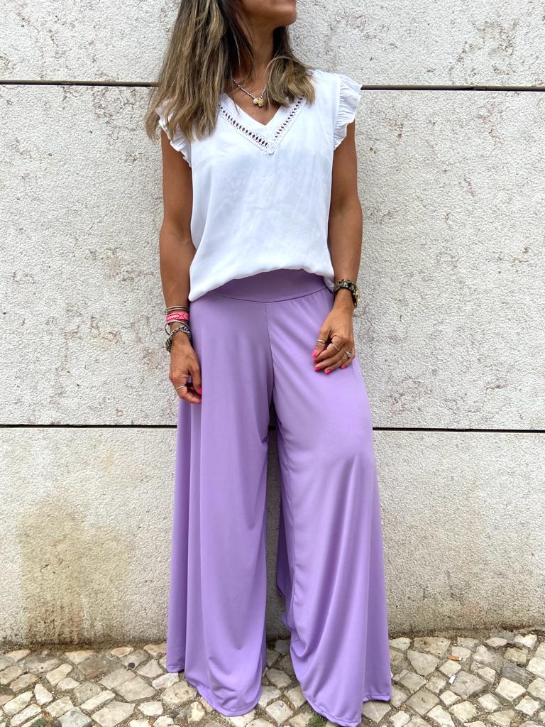 Mulher a usar calças largas lilás e blusa branca em fundo de parede de pedra clara