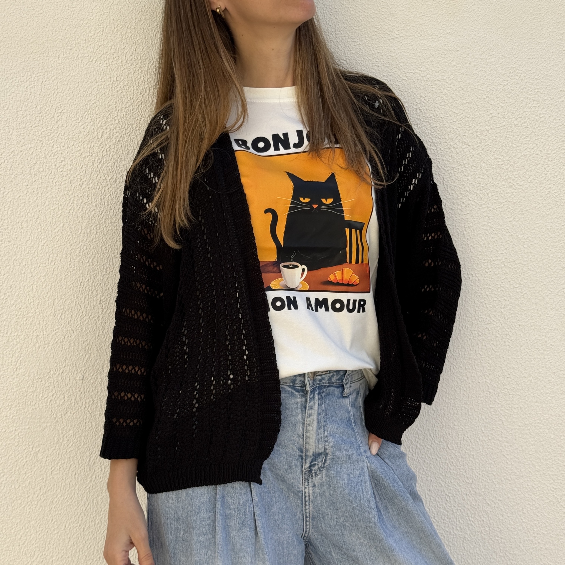 Mulher com camisola branca com gato e texto, casaco preto de malha e calças de ganga azuis.