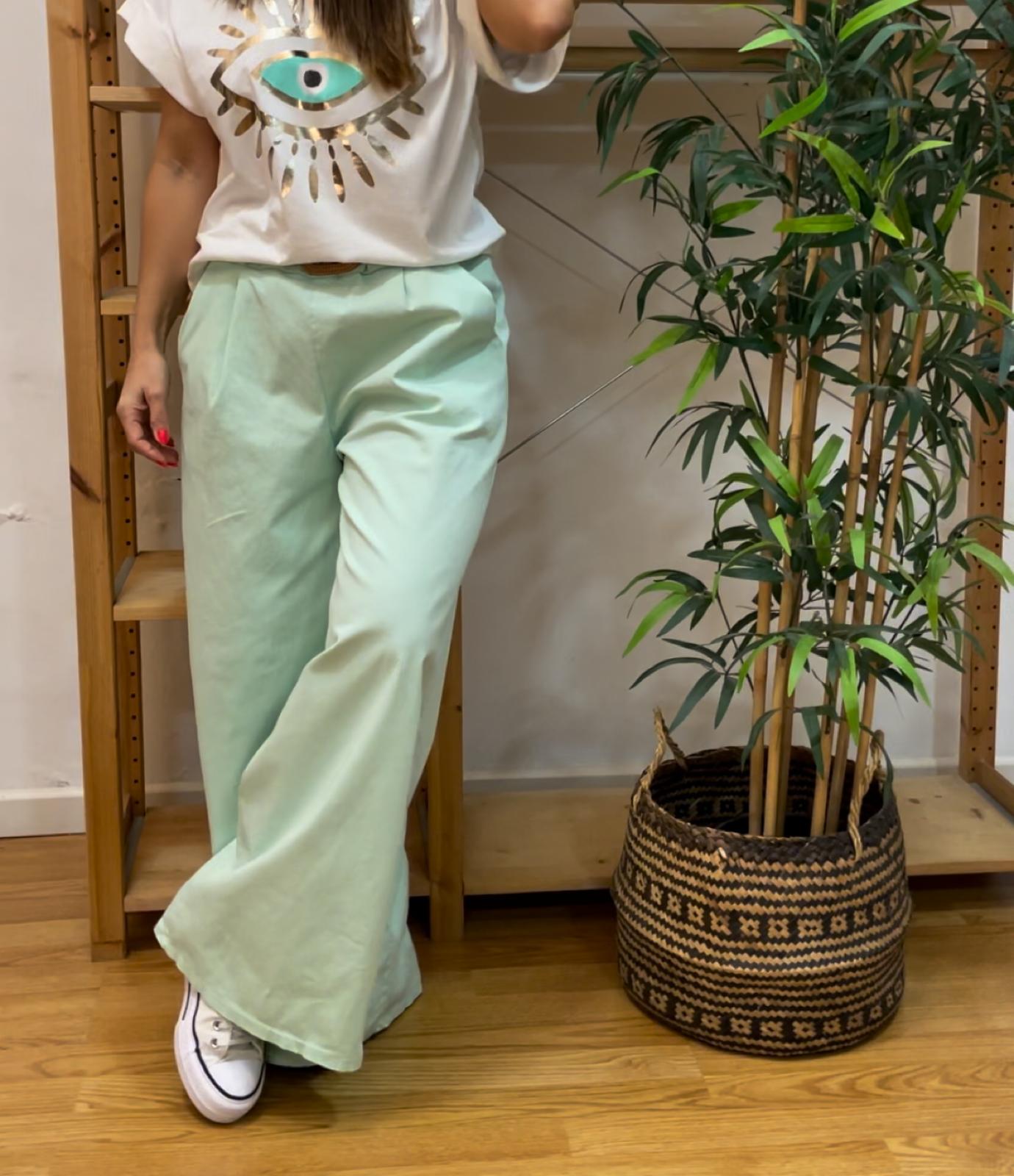 Calças verdes largas usadas com sapatilhas brancas e t-shirt com olho azul e dourado, perto de planta em vaso decorativo.