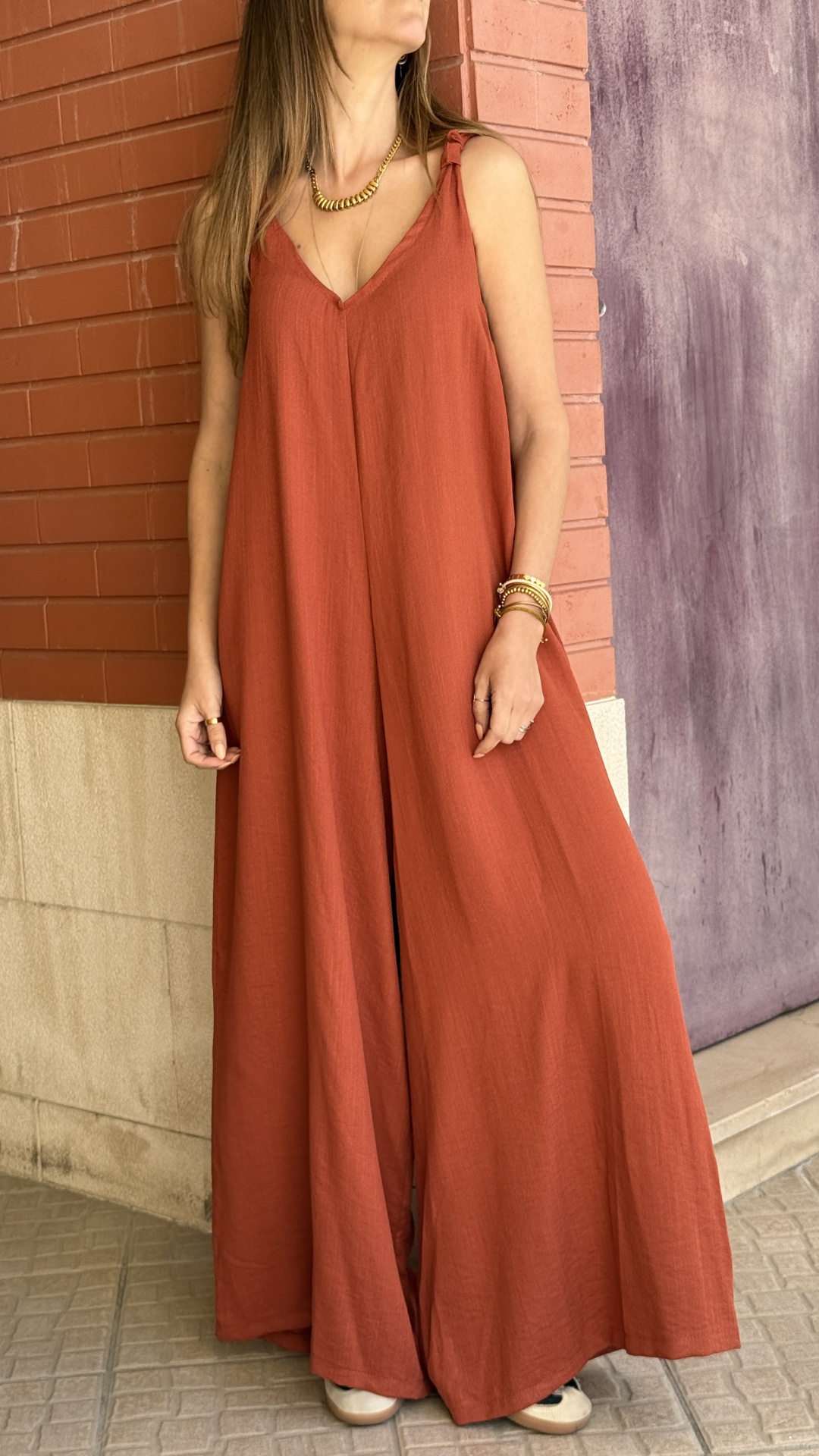 Macacão terracota largo com alças de laço, usado por mulher com joias, parede de tijolos vermelhos ao fundo