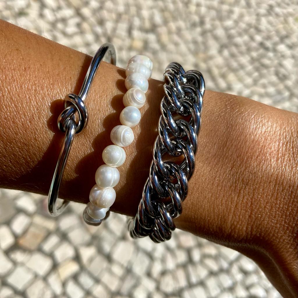 Três braçadeiras femininas em metal prateado, pérolas brancas e elos metálicos grossos usadas no braço.