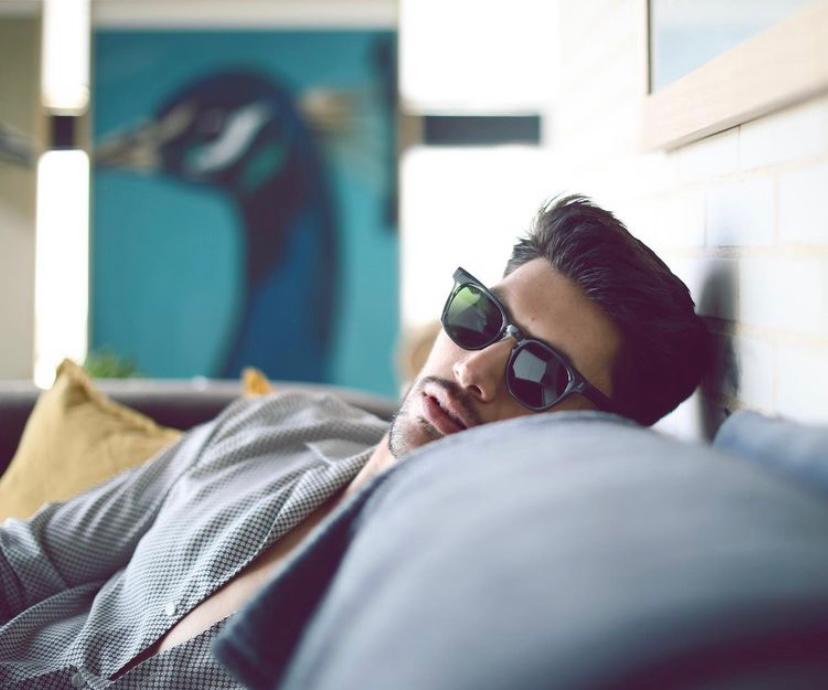 Homem de óculos de sol deitado num sofá com almofada amarela e quadro de pássaro azul
