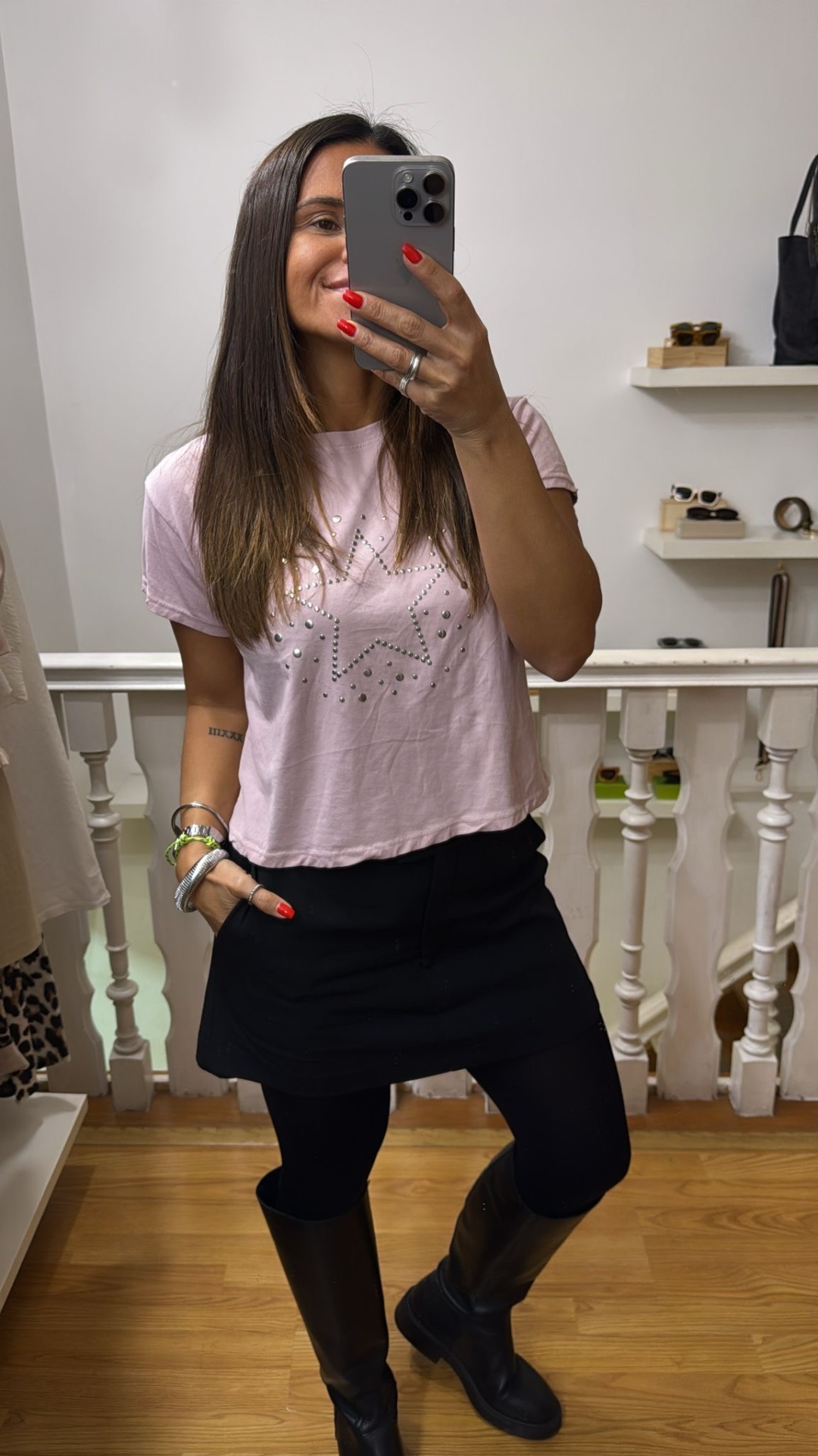 Mulher com t-shirt rosa clara com estrela de strass, saia preta, leggings e botas pretas em ambiente interior.