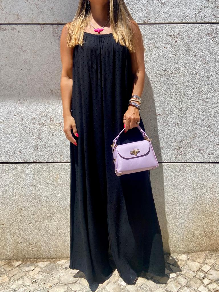 Pessoa com macacão preto e mala lilás segurando frente a parede cinza