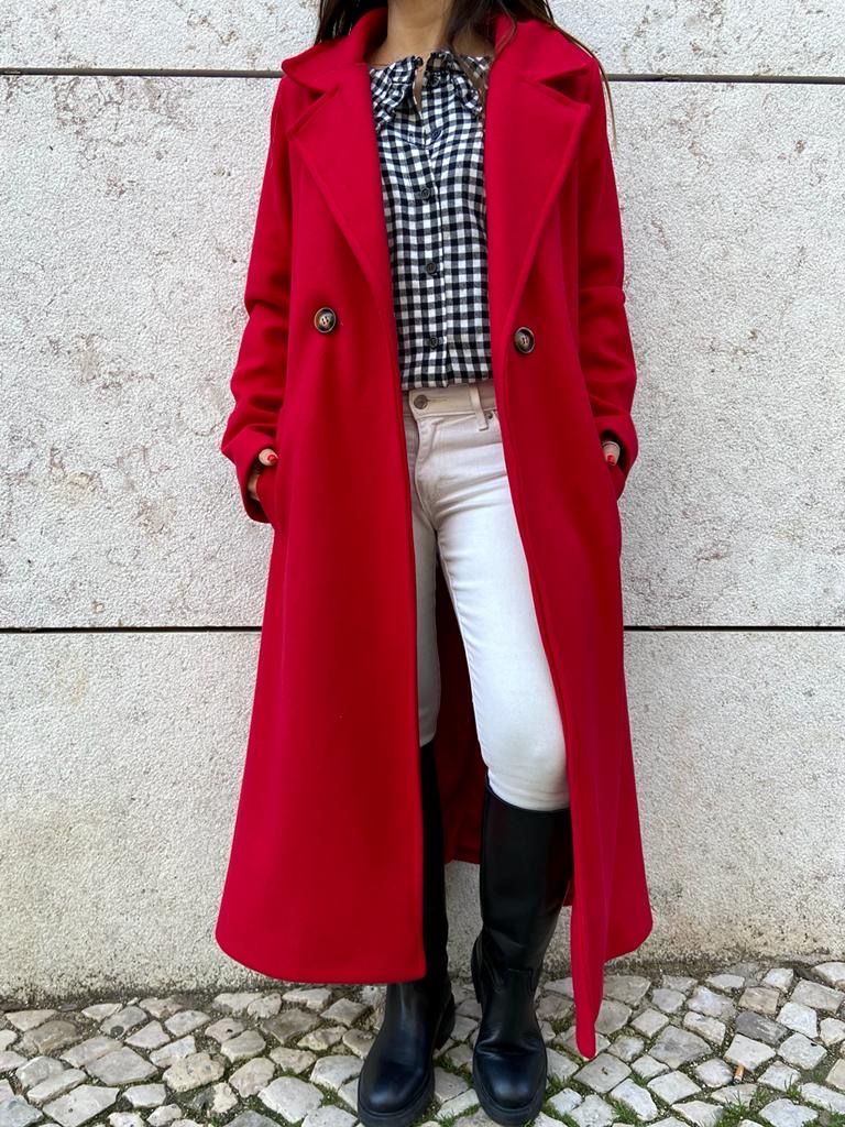 Casaco longo vermelho, camisa xadrez preta e branca, calças brancas e botas pretas em parede clara