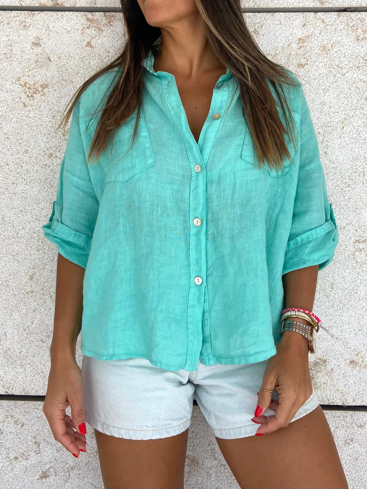 Blusa azul-turquesa leve com botões e bolsos, calções brancos e pulseiras em braço, fundo parede clara