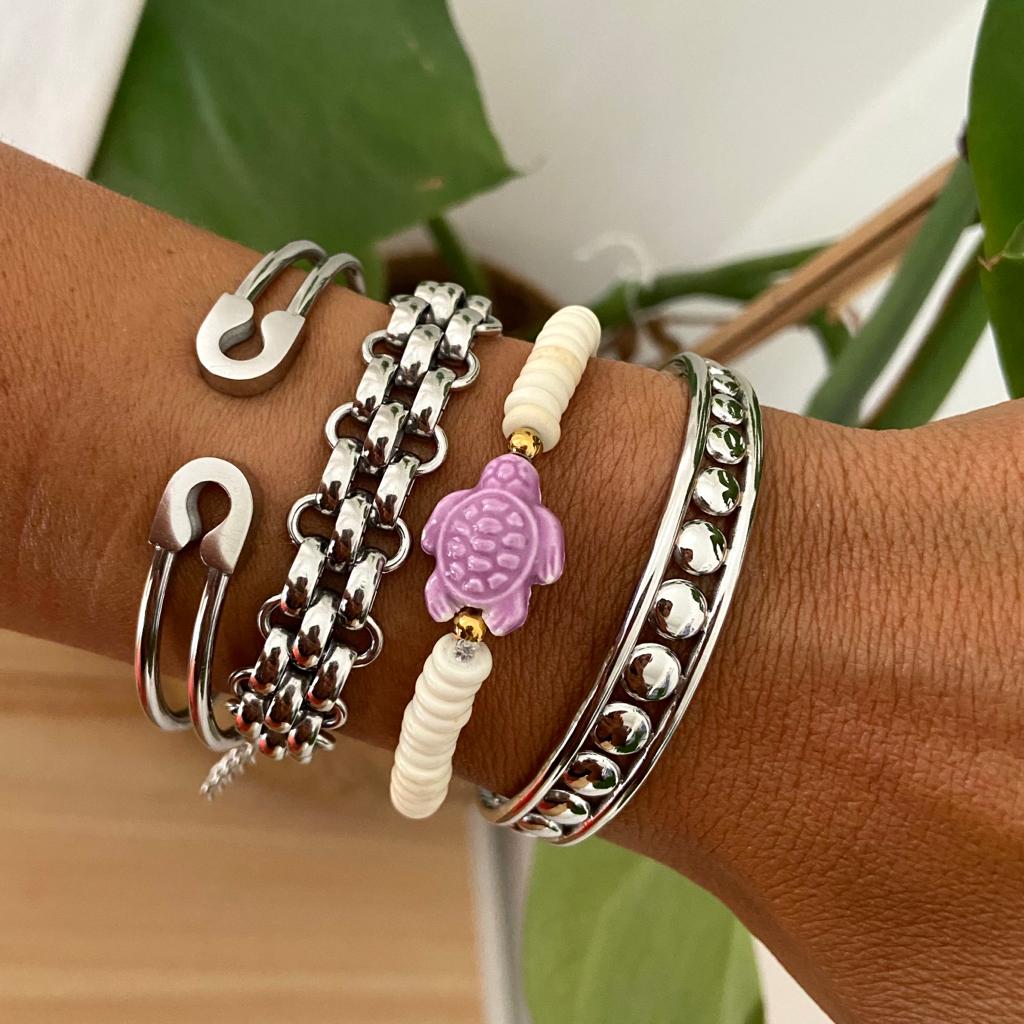 Conjunto de quatro braceletes no pulso de uma pessoa, com plantas ao fundo.