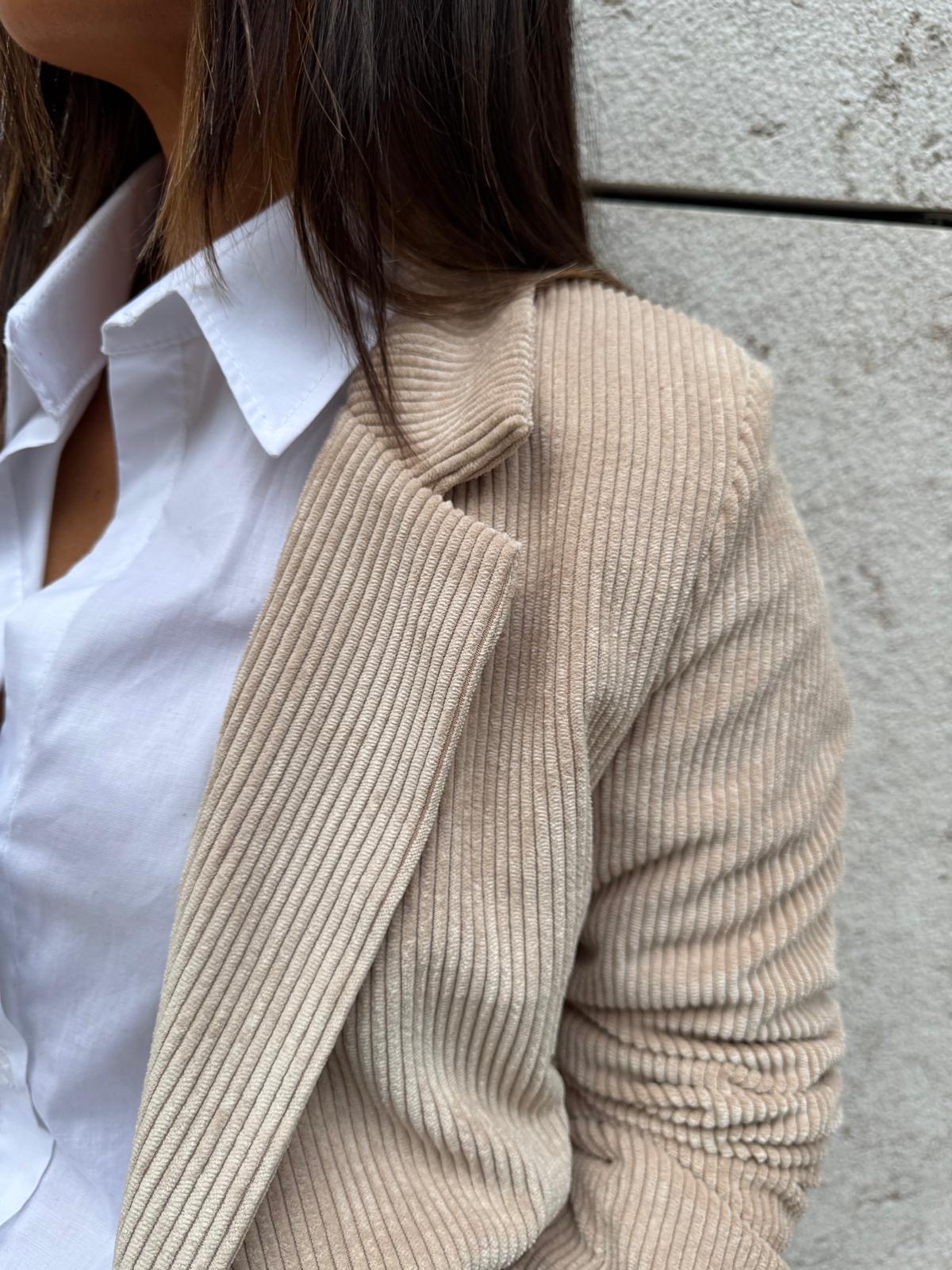 Mulher vestindo camisa branca e blazer bege canelado junto a parede de pedra clara