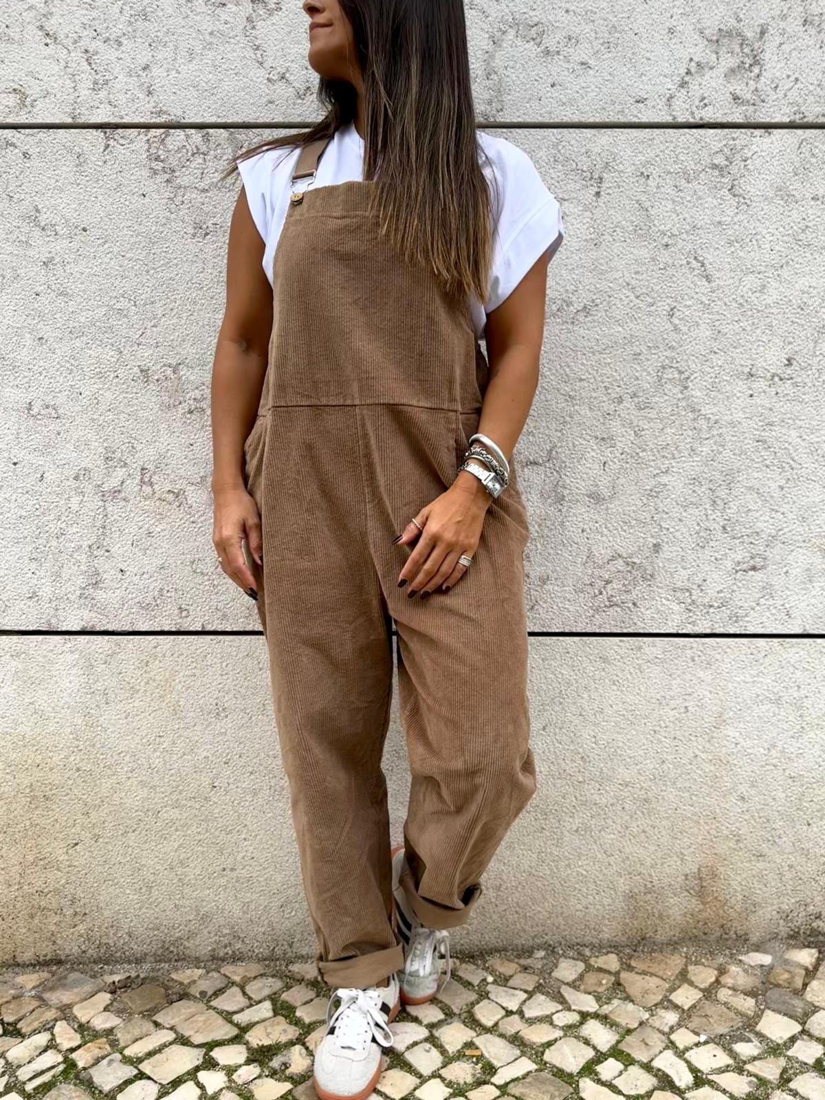 Macacão castanho de veludo com bolso frontal e alças, t-shirt branca e ténis brancos em parede de pedra.