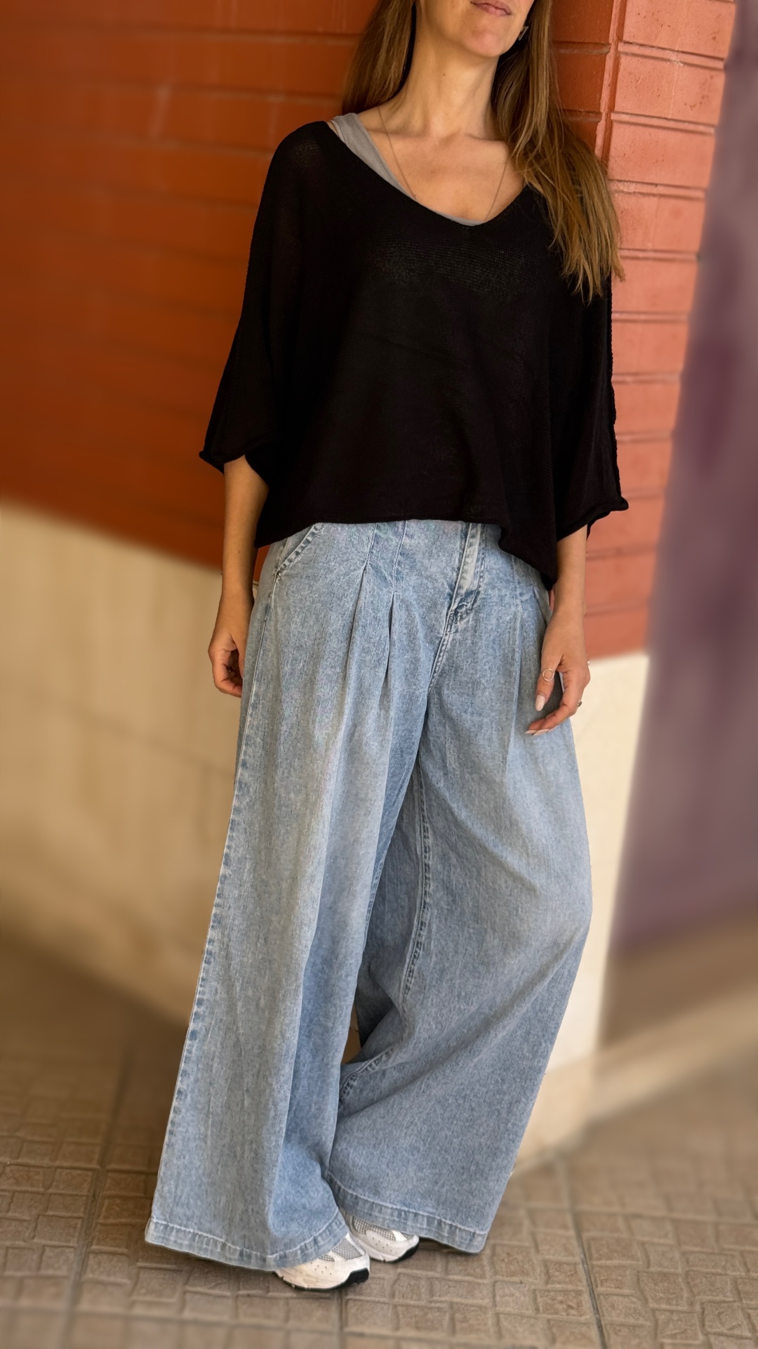 Mulher com blusa preta, calças largas de ganga e sapatilhas brancas encostada em parede de tijolos vermelhos