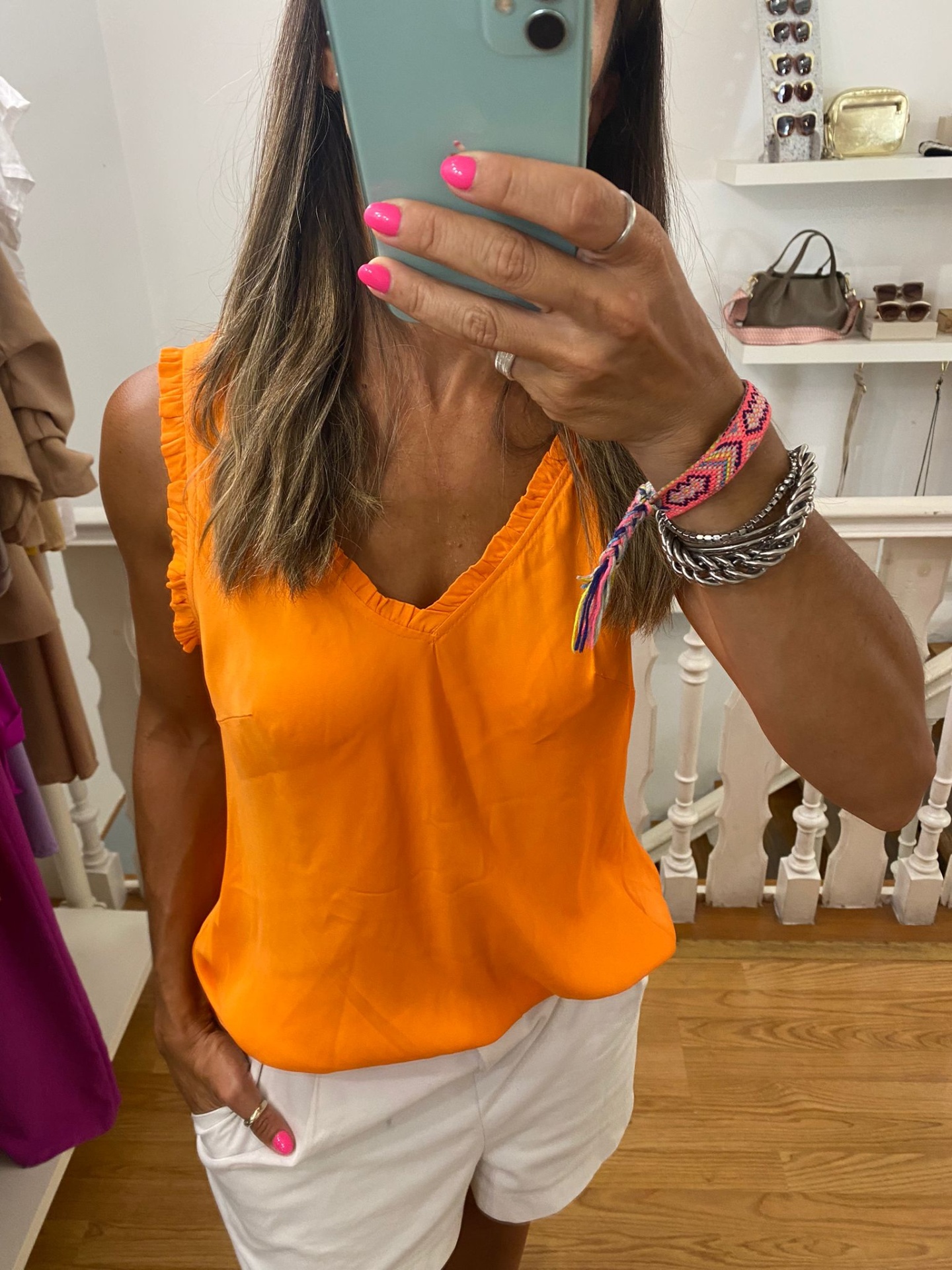 Top feminino laranja com detalhes franzidos e calções brancos, pulseiras no pulso, selfie no espelho num ambiente interior