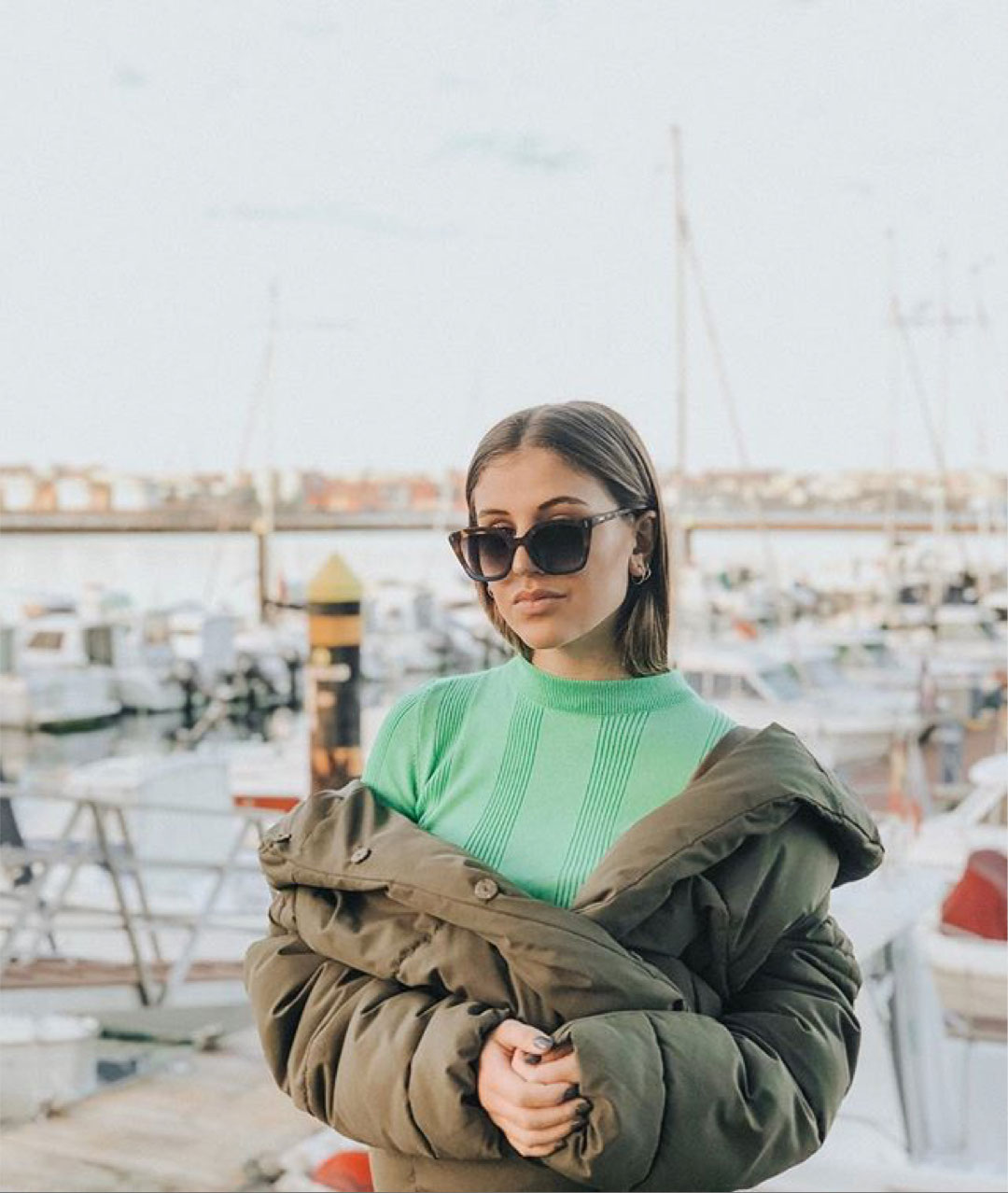 Mulher com camisola verde e casaco acolchoado junto a um cais com barcos