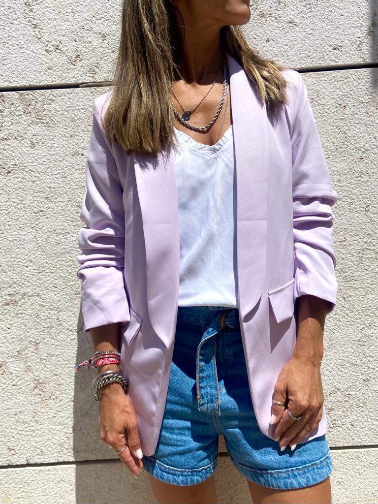Mulher com blazer lilás, t-shirt branca e calções de ganga azuis em fundo de parede clara