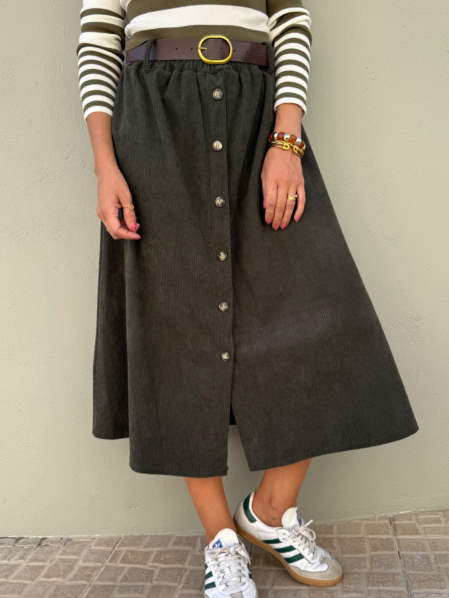 Saia midi verde-escuro em veludo com botões, usada com cinto castanho, camiseta de riscas, pulseiras e sapatilhas brancas e verdes.