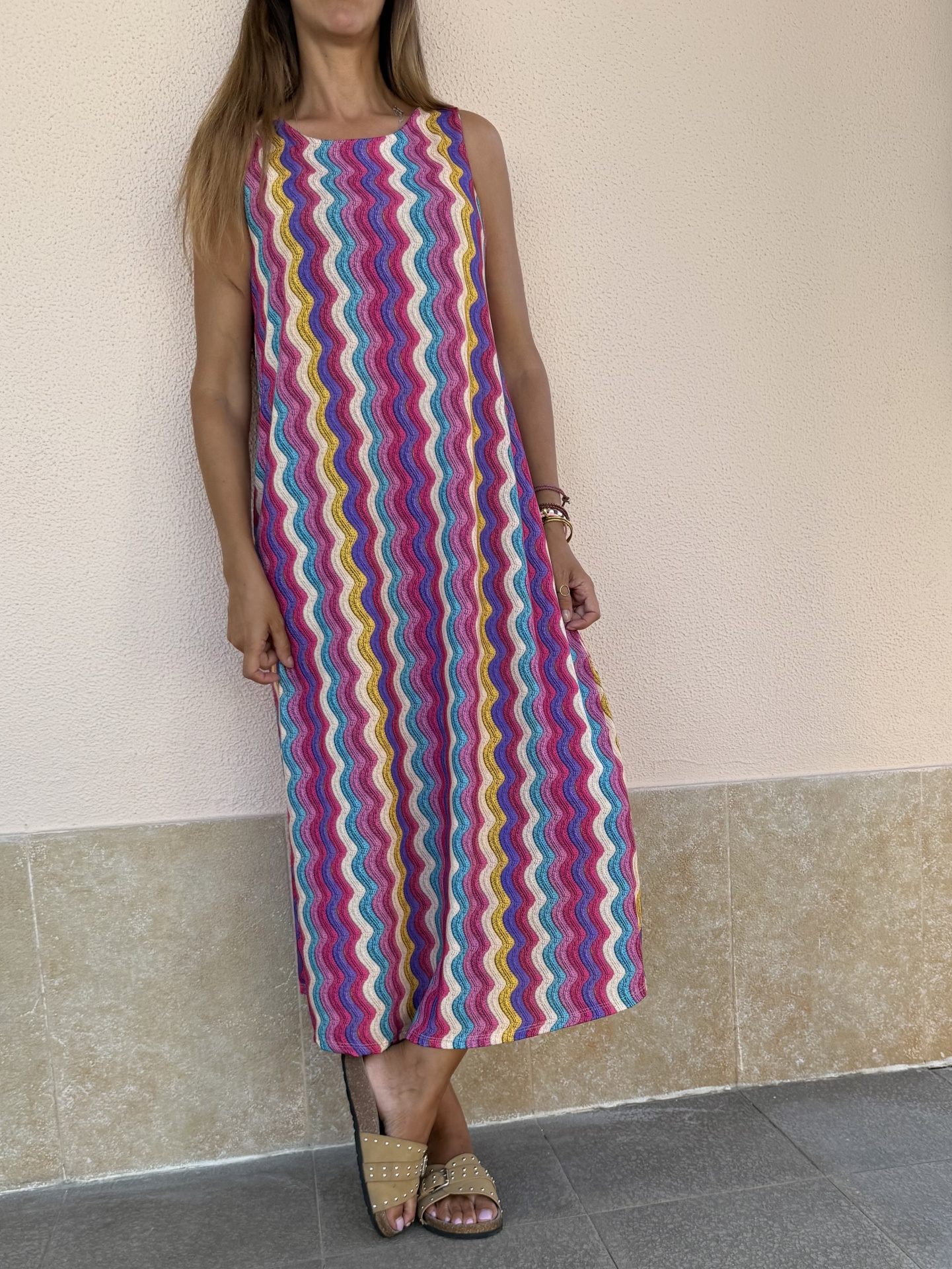 Vestido comprido com padrão de ondas coloridas e sandálias castanhas em ambiente exterior com parede clara e chão em pedra