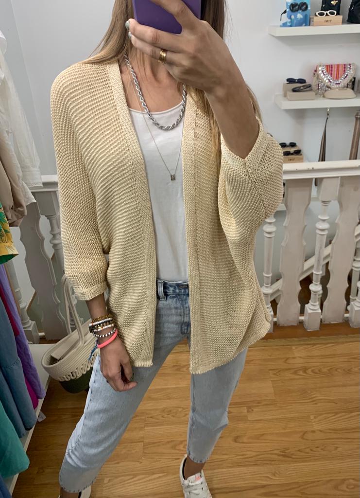 Mulher a vestir casaco de malha bege claro, t-shirt branca, jeans azuis e ténis brancos num ambiente interior