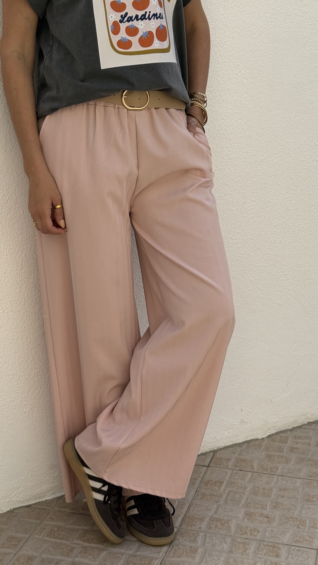 Calças largas rosa, cinto bege, t-shirt cinza com estampa, tênis pretos, piso ladrilhado e parede branca