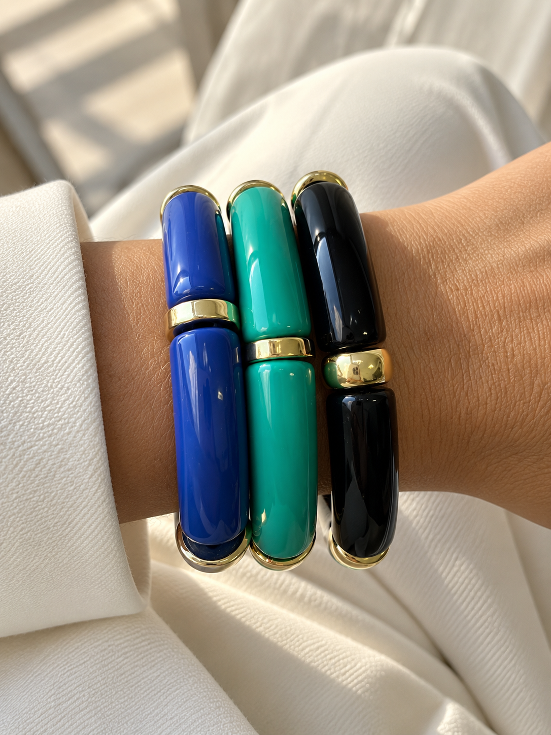 Três pulseiras brilhantes azul, verde e preto com detalhes dourados no pulso de uma pessoa.