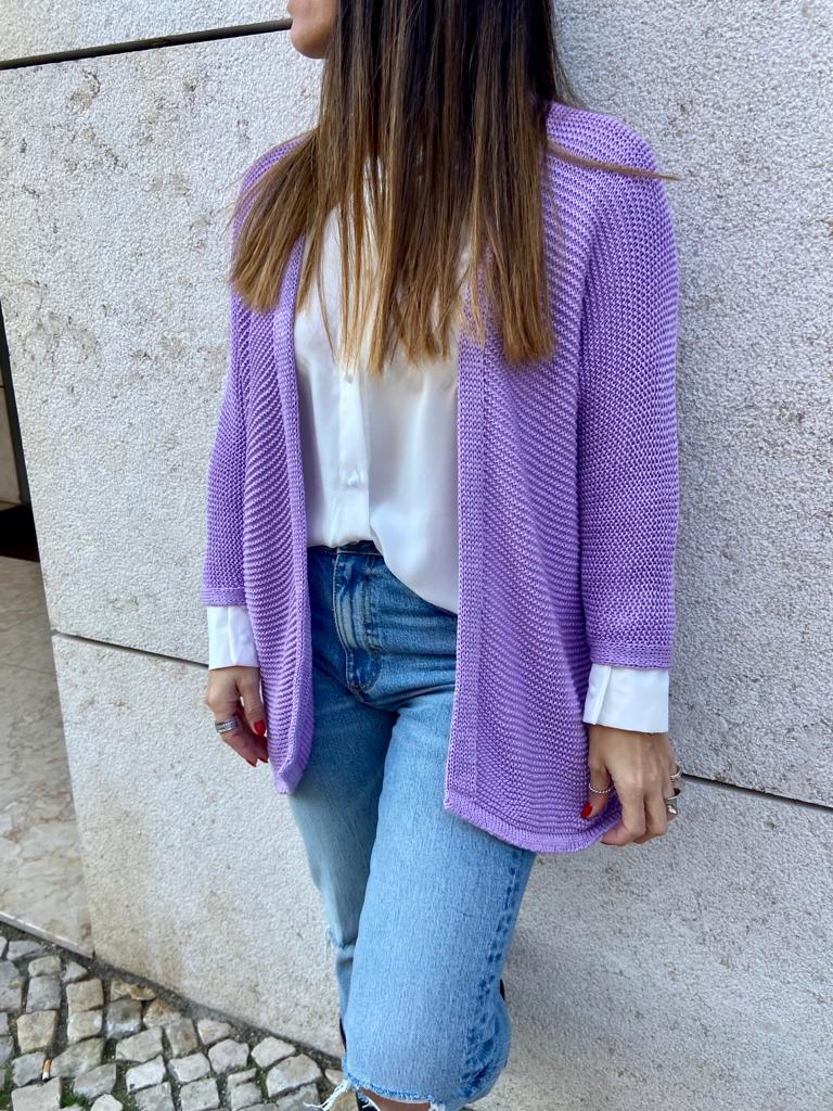 Mulher usando casaco de malha roxo sobre camisa branca e calças de ganga, parede de pedra clara ao fundo