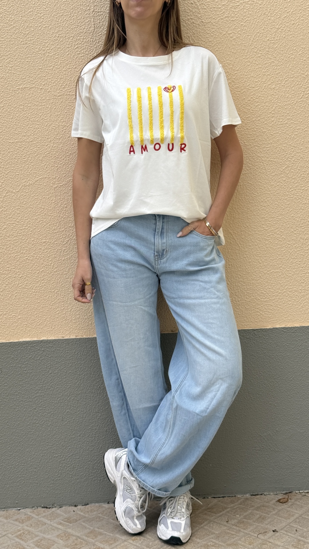 Pessoa vestindo t-shirt branca com barras amarelas e texto AMOUR, jeans azul claro e ténis brancos junto a parede bicolor