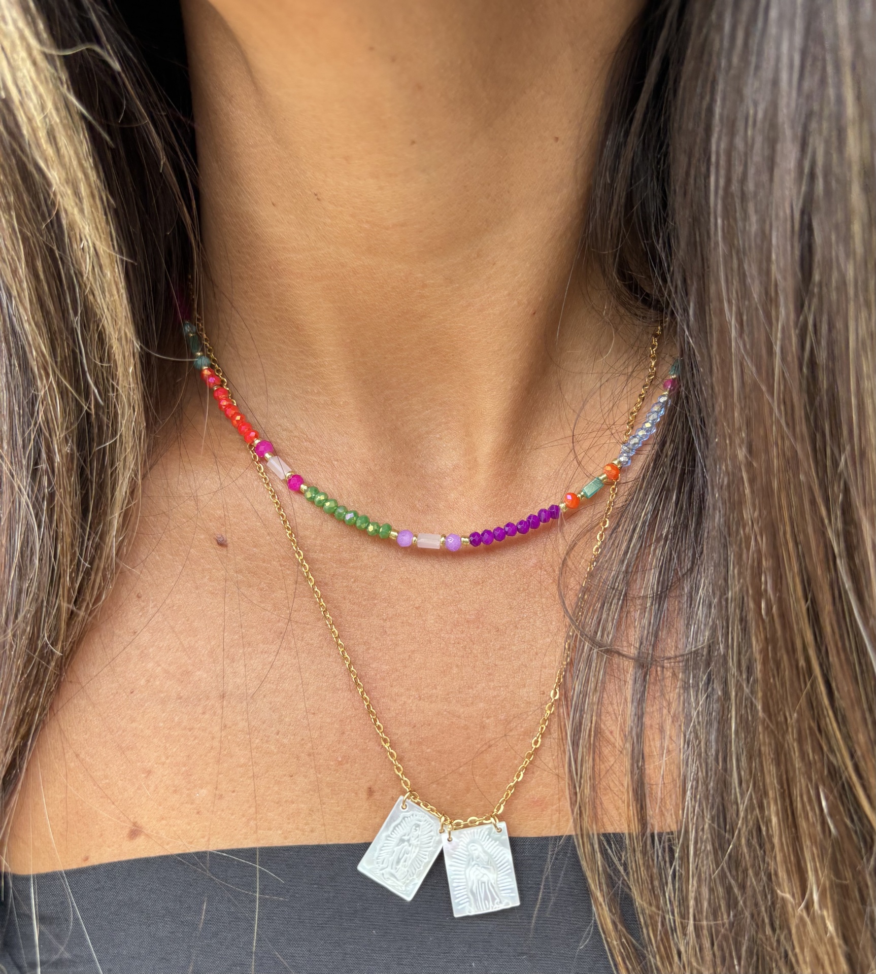 Dois colares usados no pescoço, um de miçangas coloridas e outro dourado com pendentes retangulares.