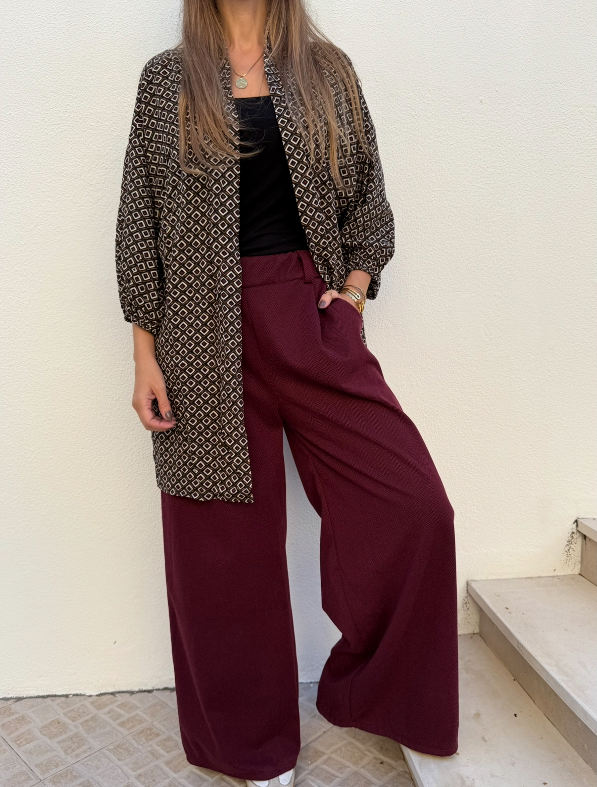 Mulher com casaco padrão geométrico castanho e branco, camisola preta e calças bordô largas