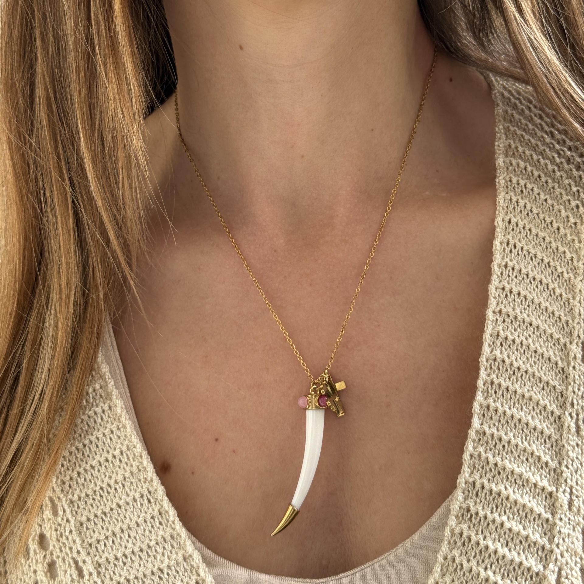 Colar dourado com pendente de dente branco e pingentes, em pescoço de pessoa com roupa bege.