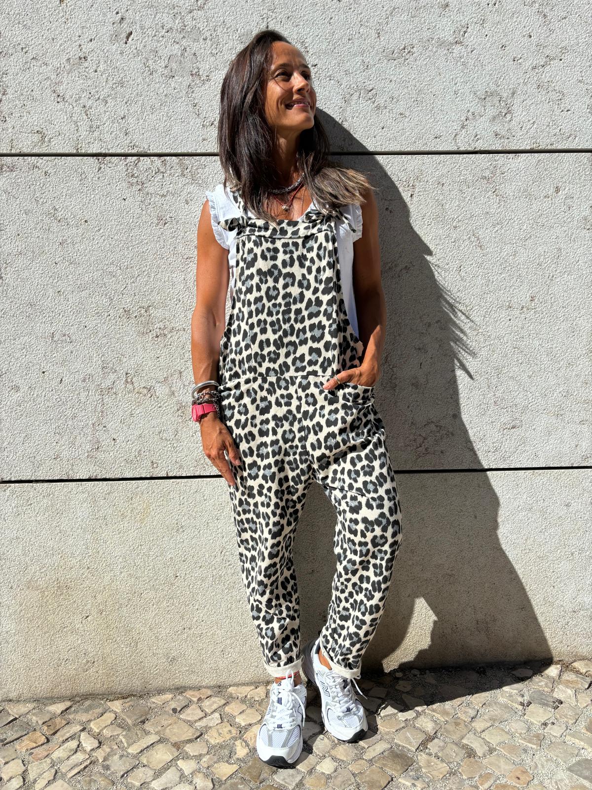 Mulher com macacão estampado animal em tecido, t-shirt branca e sapatilhas brancas, parede de pedra ao fundo.