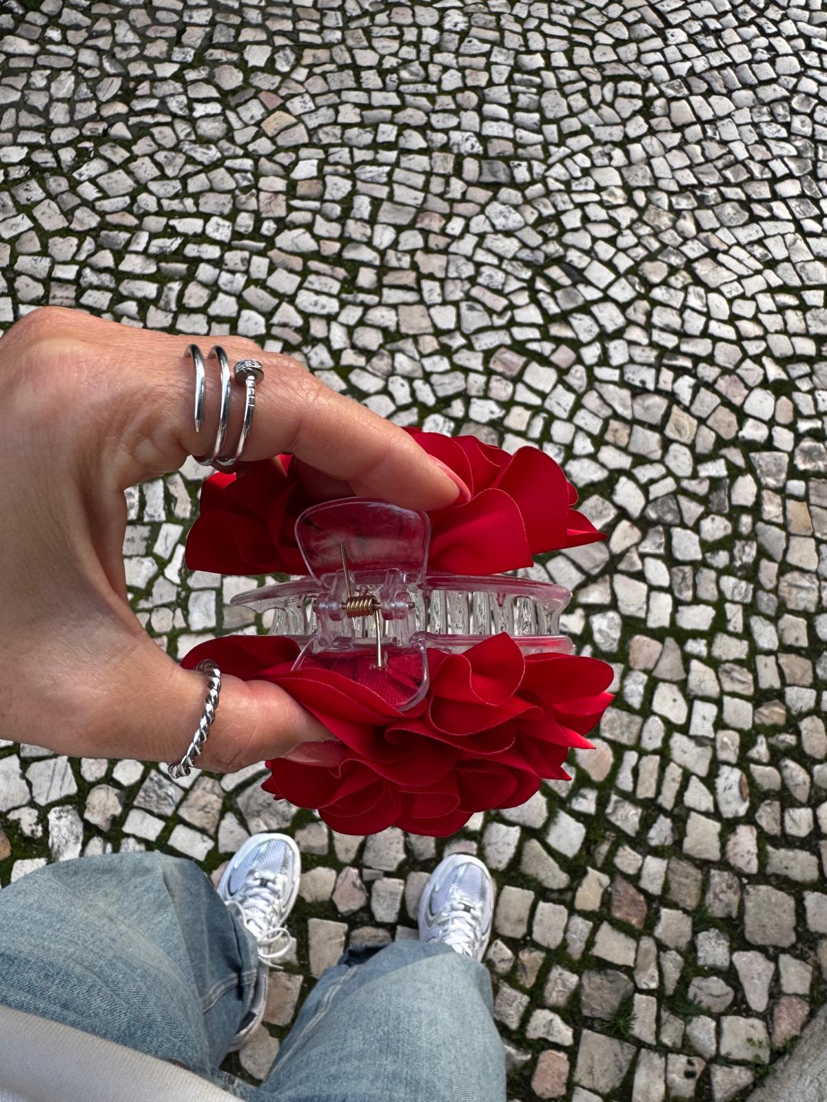 Acessório de cabelo vermelho com presilha transparente segurado por mão com anéis, sobre calçada portuguesa
