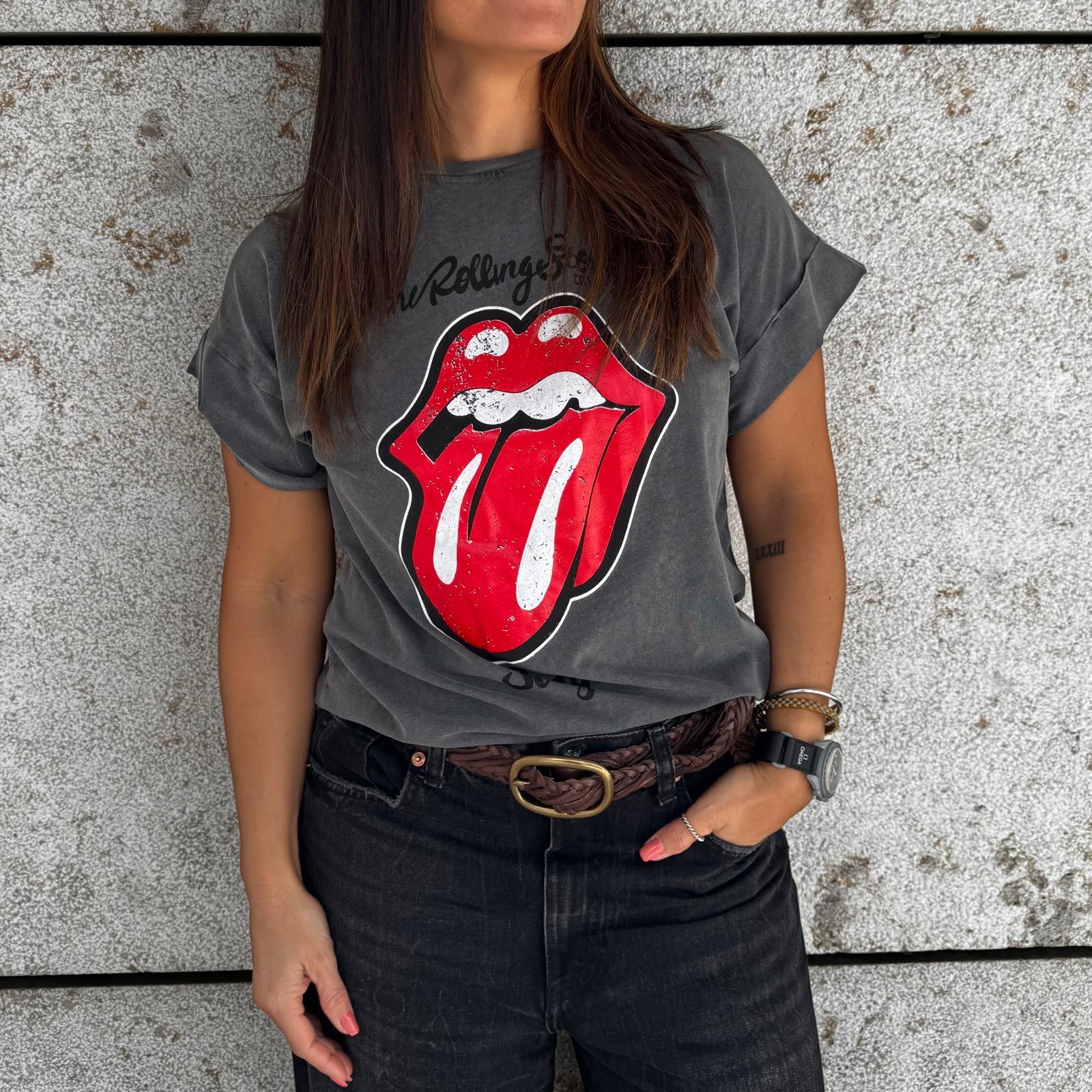 Mulher com t-shirt cinzenta The Rolling Stones, calças pretas e parede de pedra cinzenta ao fundo