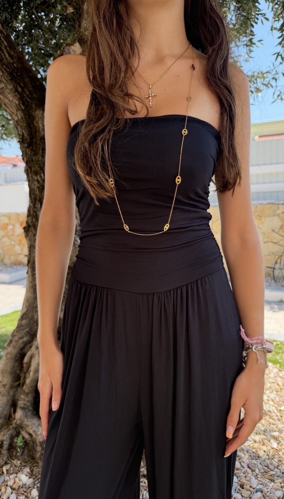 Mulher com vestido preto e colares dourados, pulseiras coloridas, em ambiente exterior com árvore ao fundo.