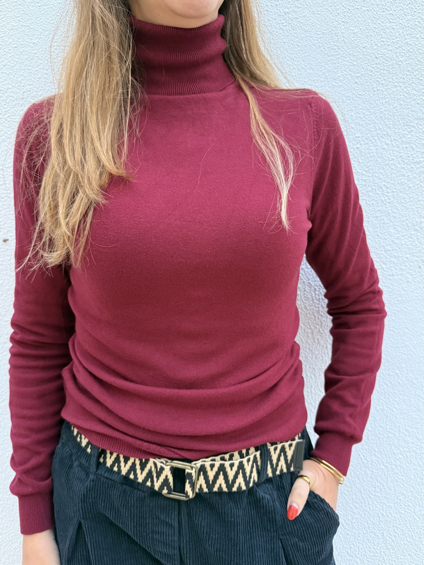 Pessoa vestindo suéter vinho de gola alta, cinto com padrão zig-zag e calças azul marinho de veludo cotelê
