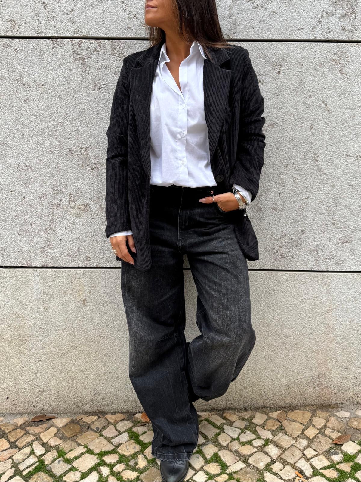 Indivíduo veste blazer preto risca de giz, camisa branca, calças pretas largas e sapatos pretos em parede cinzenta