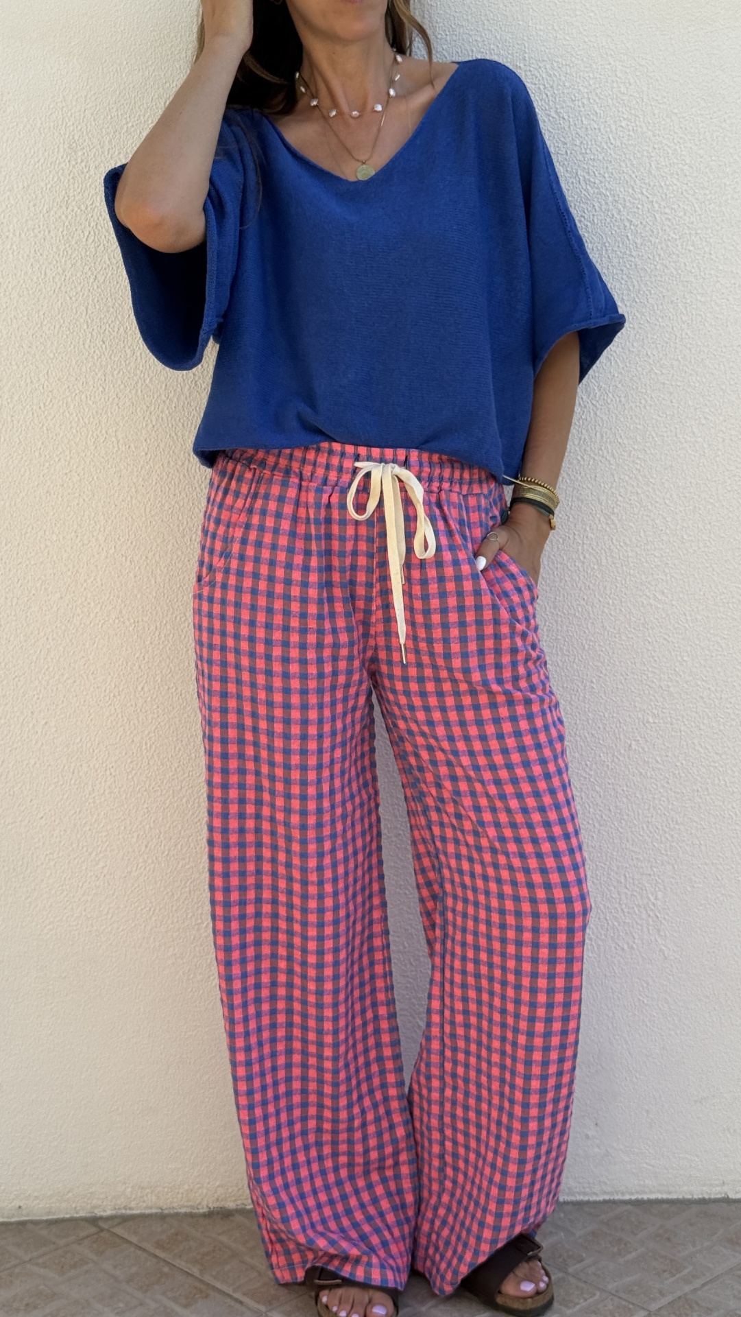 Pessoa com t-shirt azul e calças largas quadriculadas em rosa e azul, usando sandálias e pulseiras, encostada a uma parede branca.