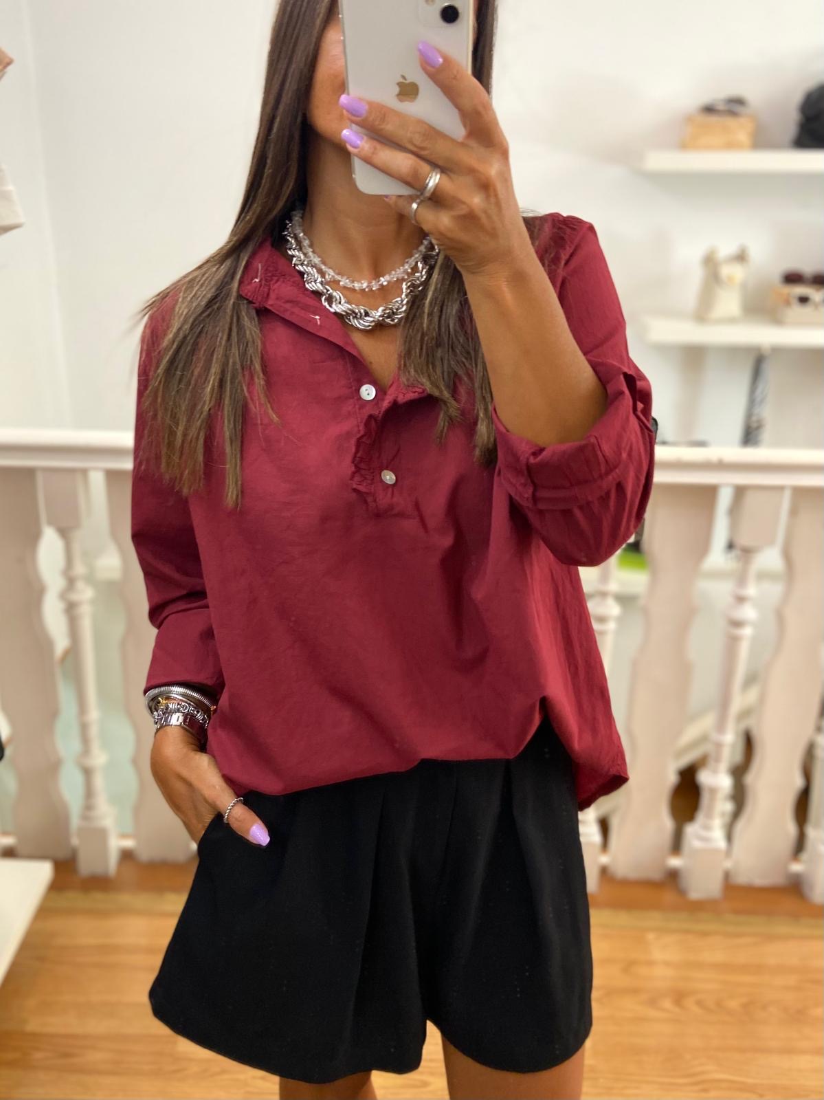 Mulher veste blusa bordô de manga comprida e calções pretos, tirando selfie com telemóvel no interior