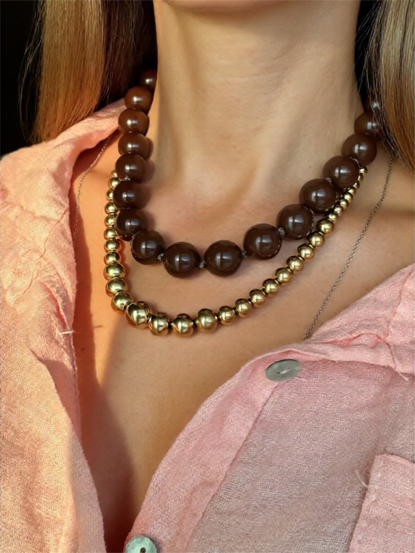 Colar duplo com contas castanhas e douradas usado sobre camisa rosa claro