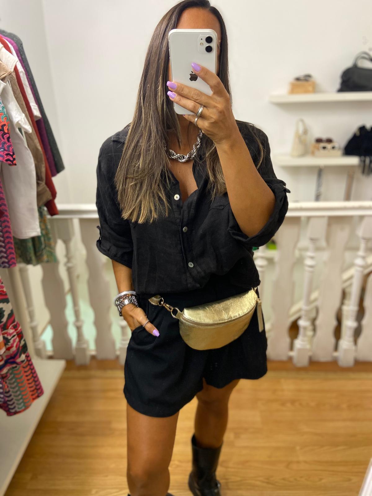 Mulher com camisa e calções pretos, botas e pochete dourada a tirar selfie num espelho