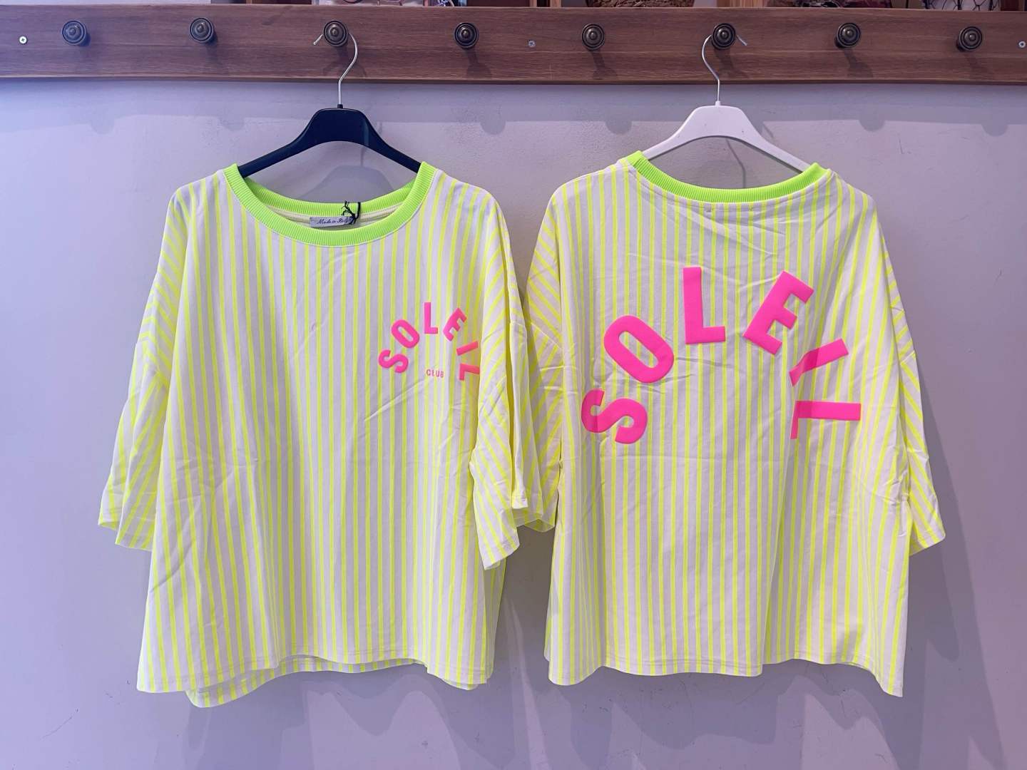 Duas t-shirts amarelas às riscas com texto cor-de-rosa penduradas na parede