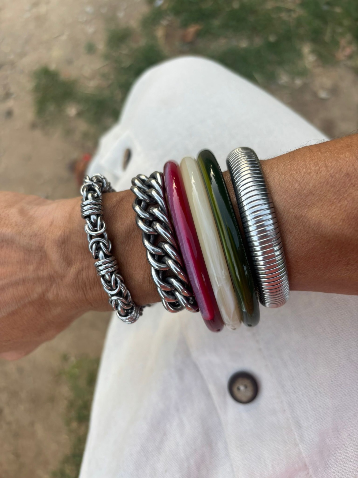 Pulso com pulseiras de metal e plástico em cores variadas, sobre calça branca e chão de terra