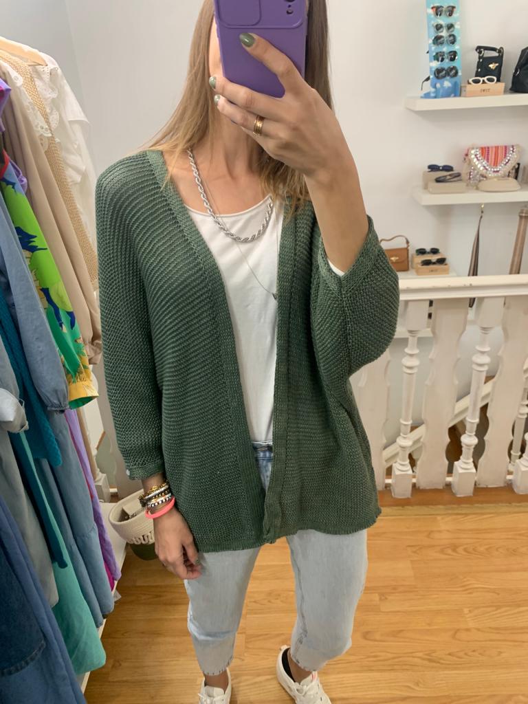 Cardigan verde de malha com riscas, t-shirt branca, calças de ganga claras num quarto com acessórios e roupas penduradas