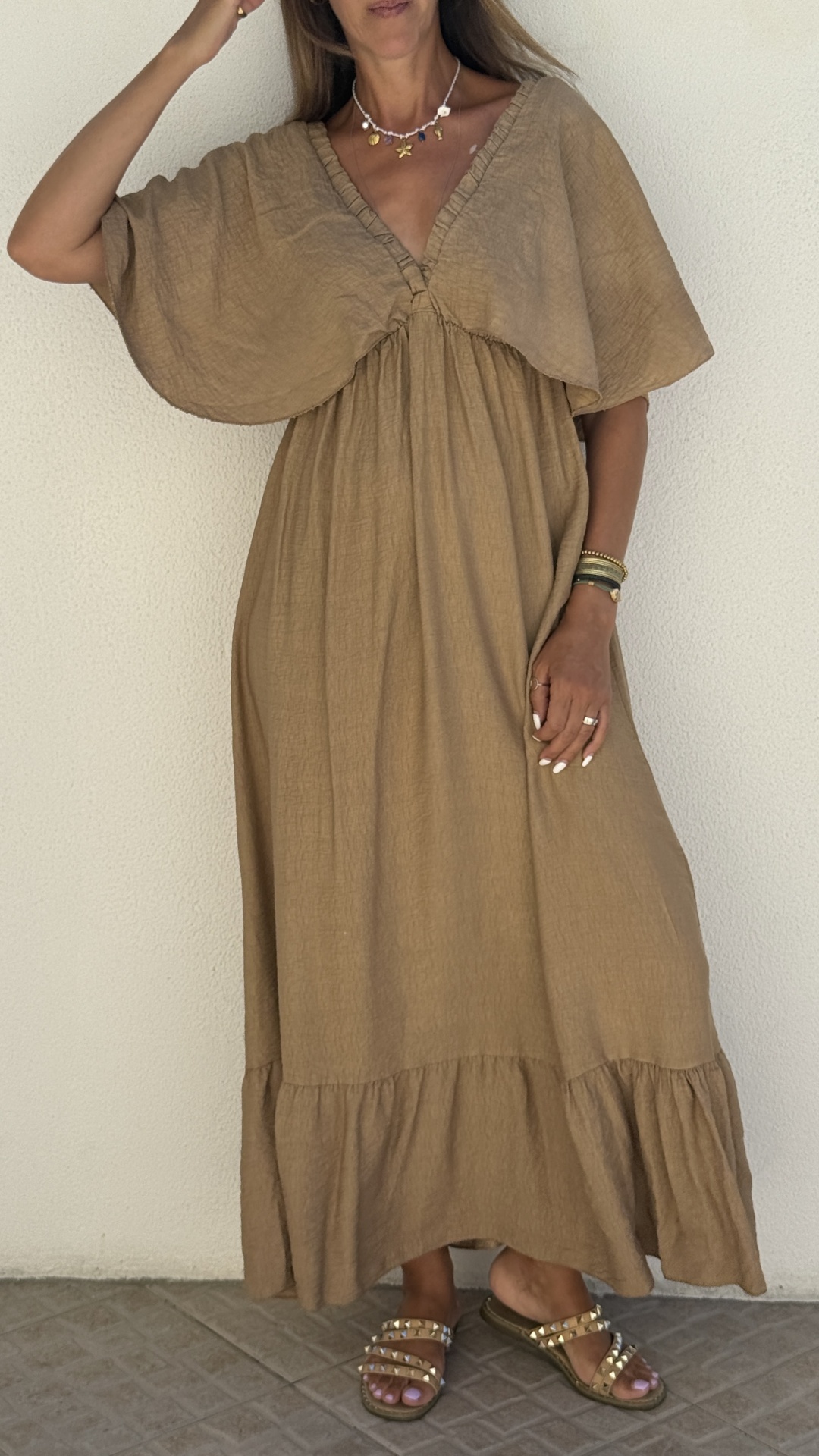Mulher com vestido comprido castanho de tecido leve e mangas amplas, com acessórios e sandálias, em fundo simples branco e chão de azulejo.
