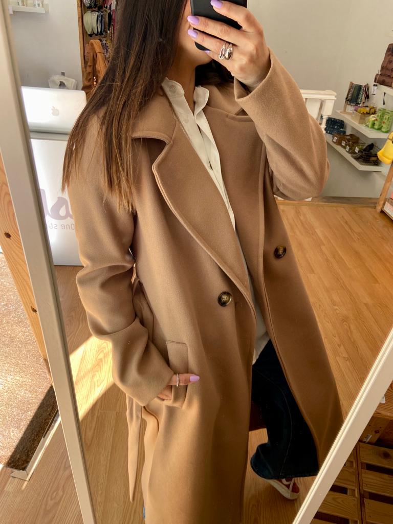 Casaco bege comprido com abotoamento duplo e camisa branca num ambiente interior com chão de madeira.