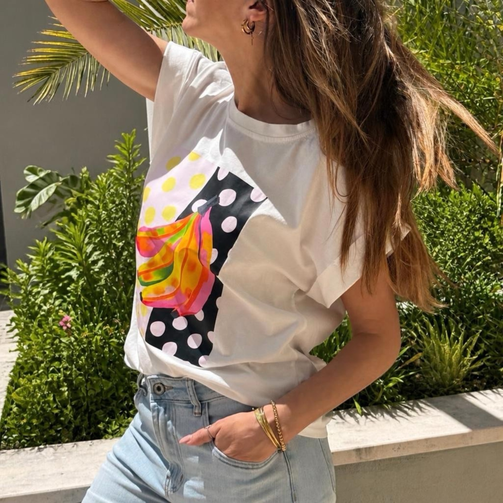 Pessoa usando t-shirt branca estampada colorida e calças de ganga azul clara em ambiente exterior com plantas.