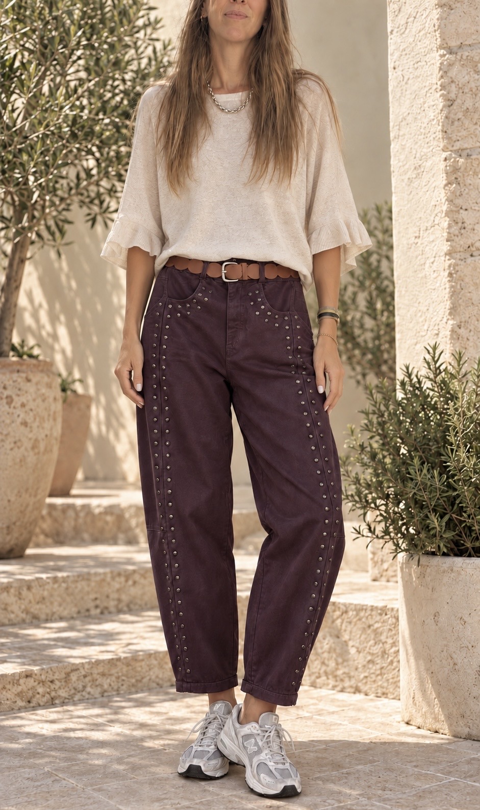 Mulher com blusa bege, calças vinho com tachas e sapatilhas branca e cinzenta ao ar livre