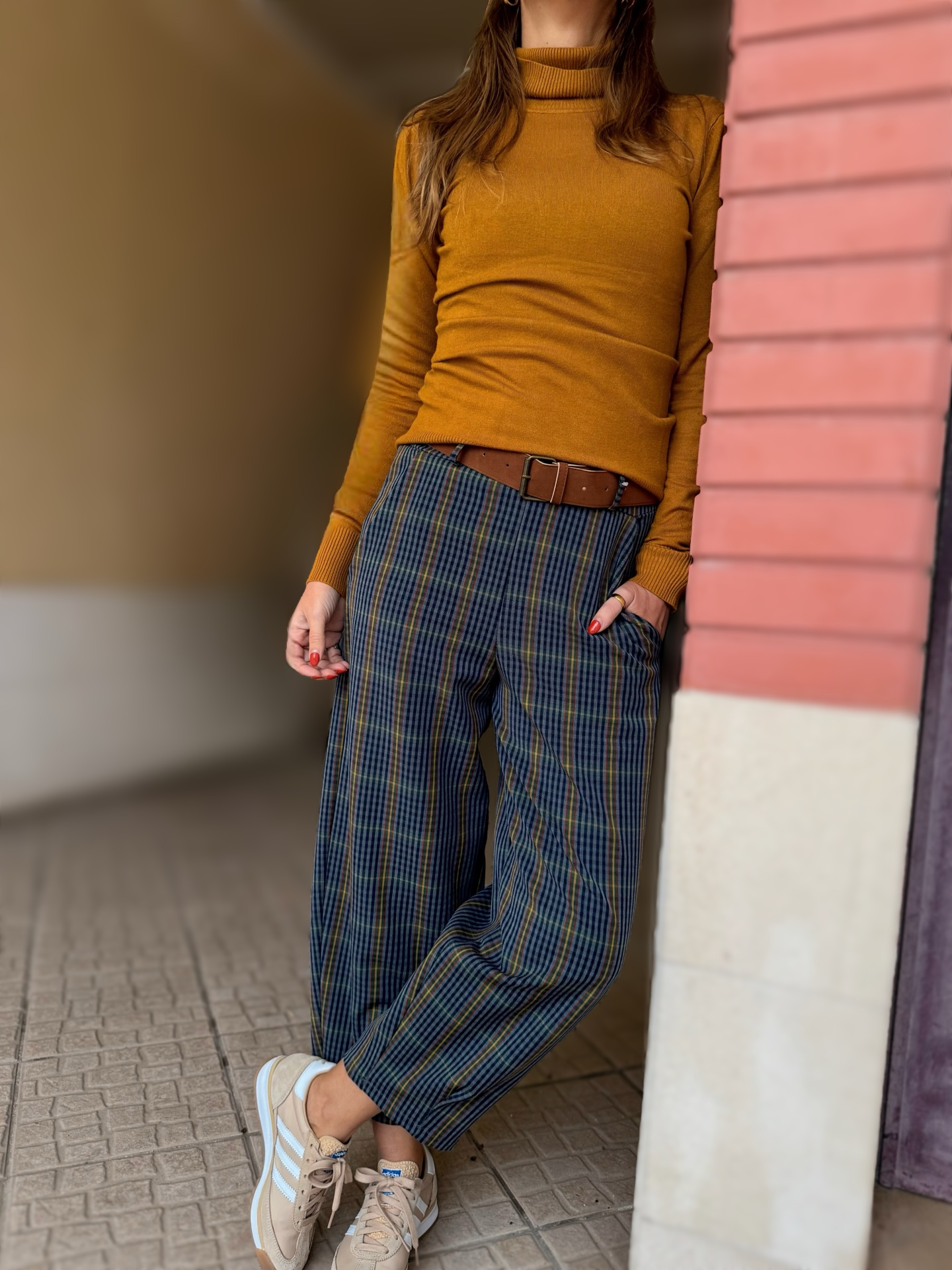 Pessoa vestindo camisola castanha, calças às riscas largas e ténis bege, encostada a parede de tijolos vermelhos.