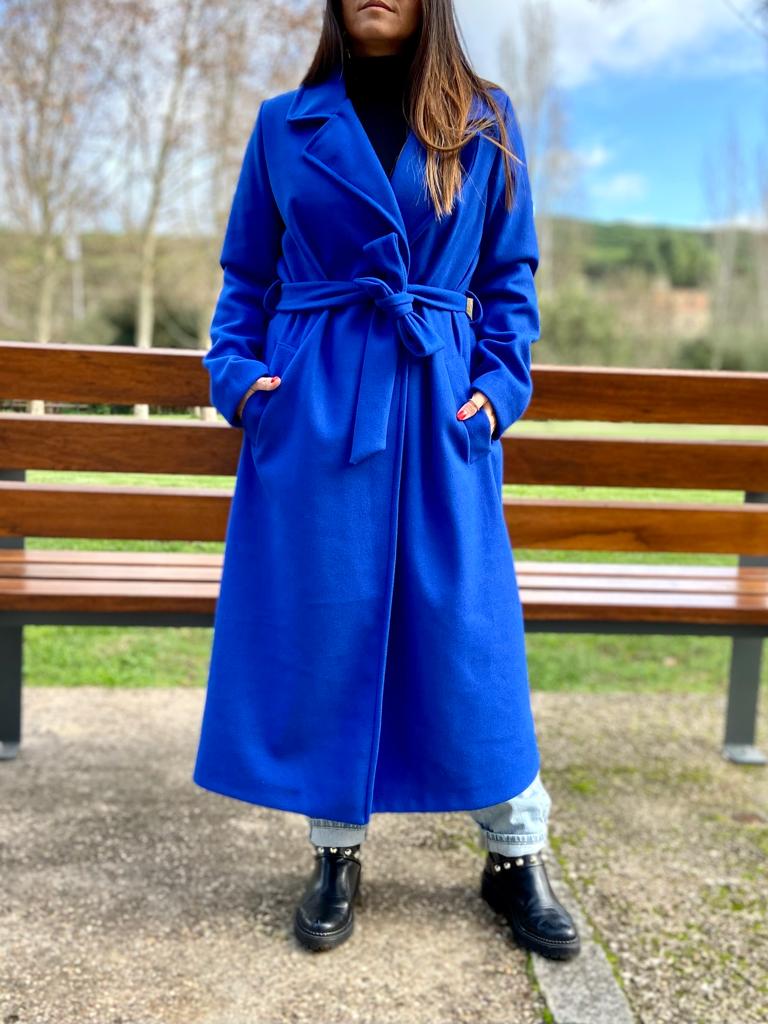 Casaco comprido azul royal com cinto e lapelas largas, pessoa de botas pretas num parque